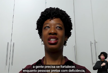 Foto de isadora dos ombros para cima, atrás dela um armário branco, ela usa gloss e camiseta vinho. Sobre a imagem a legenda: “Nós precisamos nos fortalecer enquanto pessoas pretas com deficiência.”. No canto inferior direito a intérprete de libras, mulher negra de cabelo black e roupas pretas.